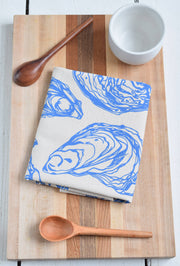 Hearth and Harrow - Oyster Tea Towel in Blue-violet - Organic Cotton displayed on a cutting board with a wooden spoon, showcasing its intricate sea shell design.
