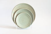 RachaelPots - Dinner Plate: Nude/Light Blue set of three mint-green speckled ceramic plates, hand-thrown and displayed nested on a clear stand against a white background.