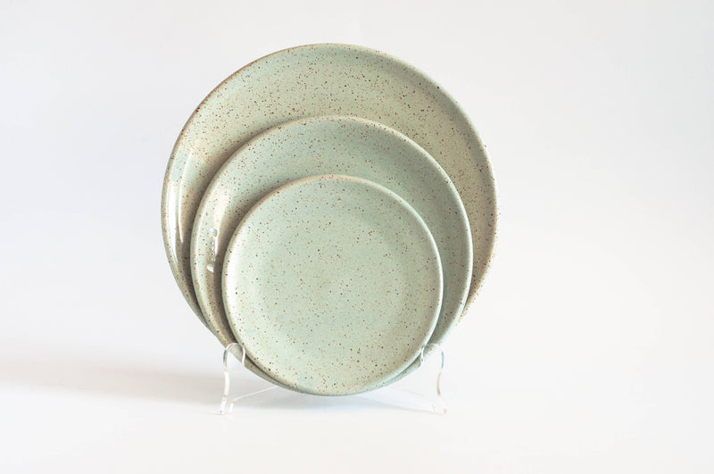 RachaelPots - Dinner Plate: Nude/Light Blue set of three mint-green speckled ceramic plates, hand-thrown and displayed nested on a clear stand against a white background.