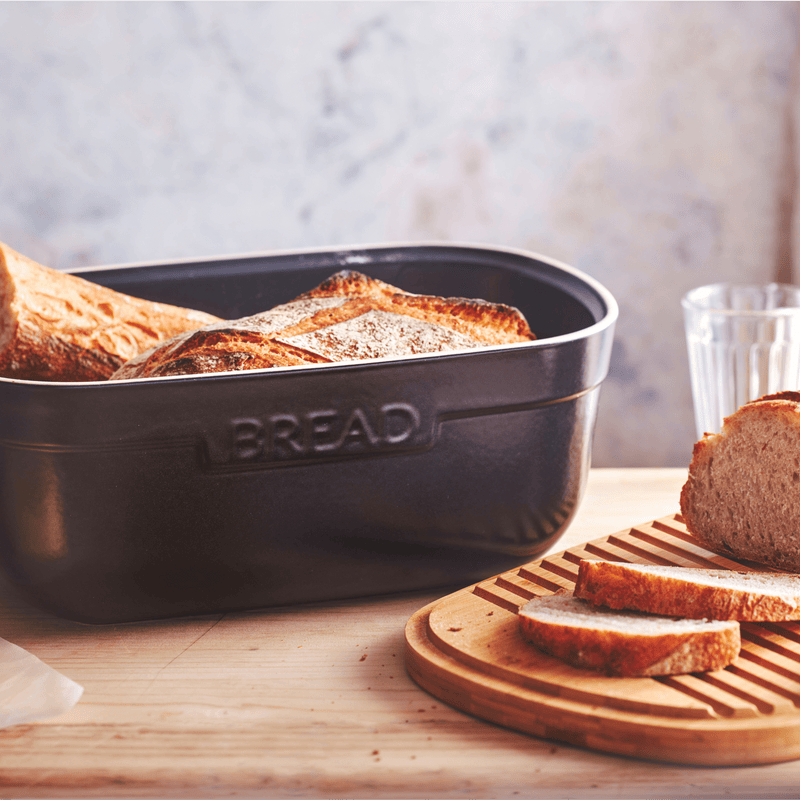 Emile Henry | Bread Box, Burgundy on a light wood countertop with sliced rustic loaf on a wooden board, showcasing its elegant ceramic design and multifunctional lid.