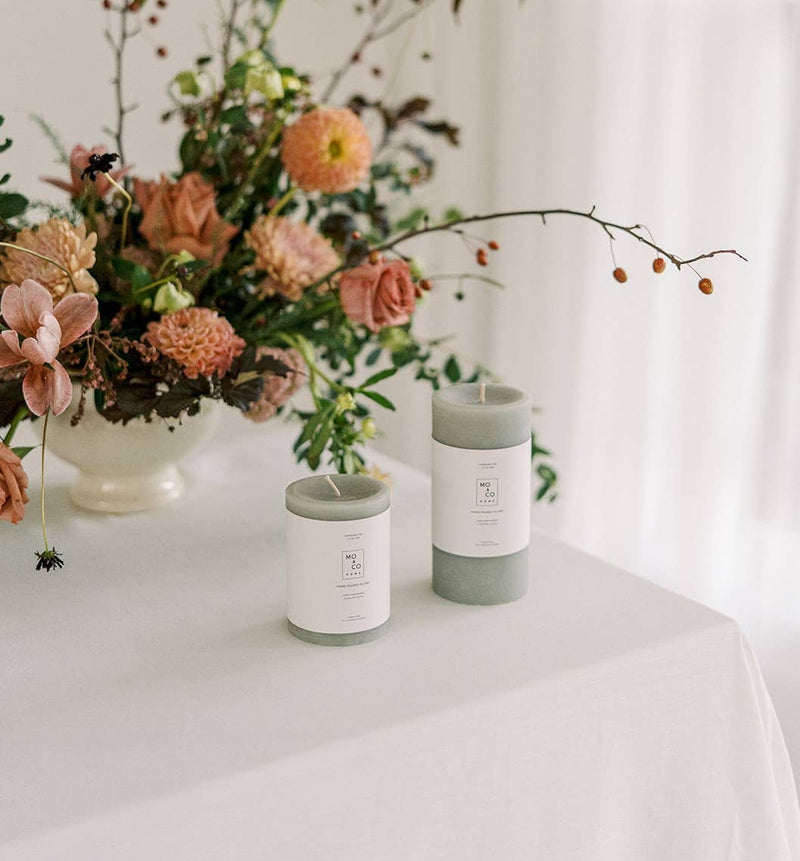 Mo&Co Home - 100% Beeswax Pillar Candles | Eucalyptus: 3x4 Inch. Two candles on a table with a floral centerpiece in a kitchen setting.