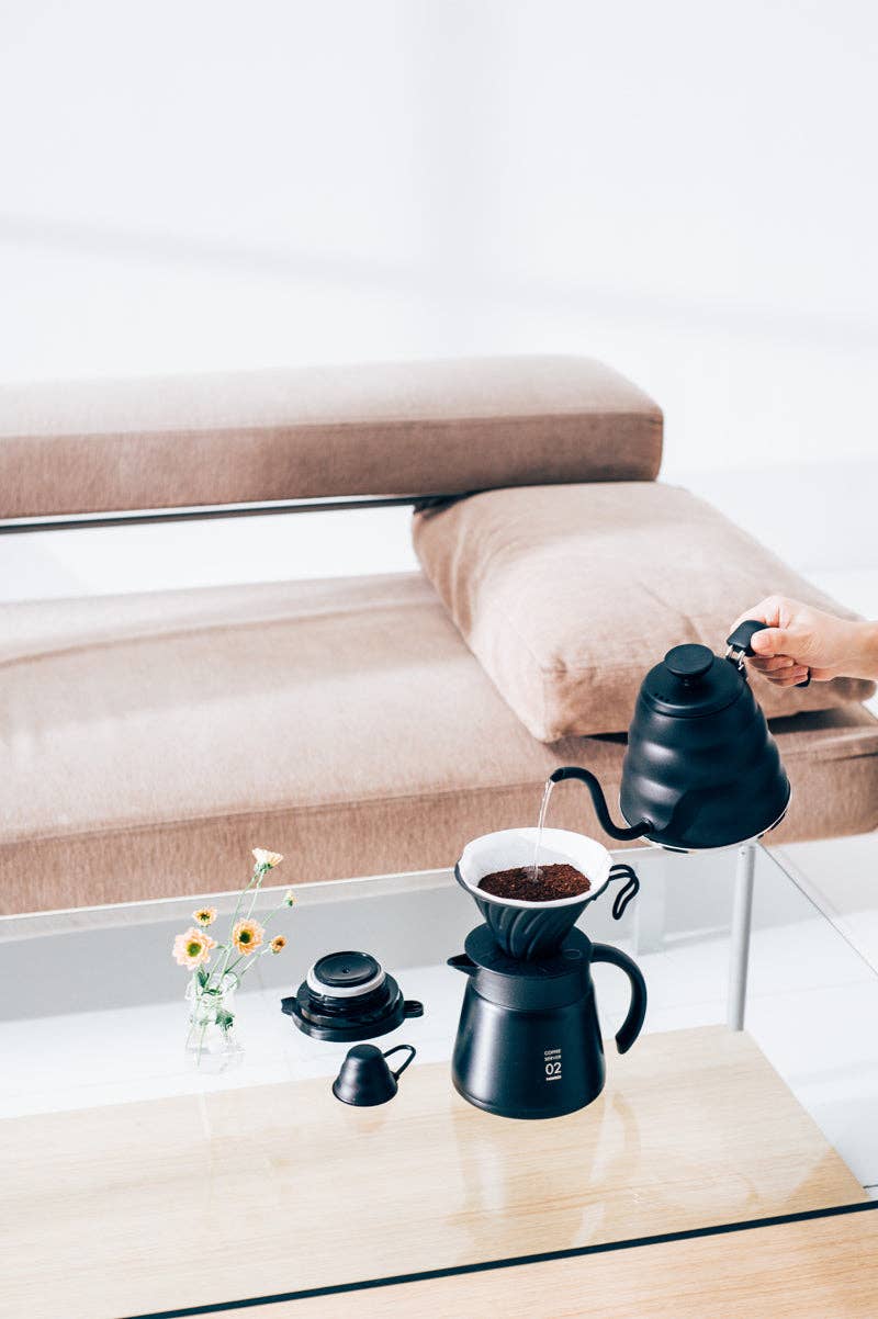 HARIO USA - V60 Metal Dripper: Silver on a glass table with a black kettle, carafe, and yellow flowers, showcasing minimalist coffee brewing in a modern setting.