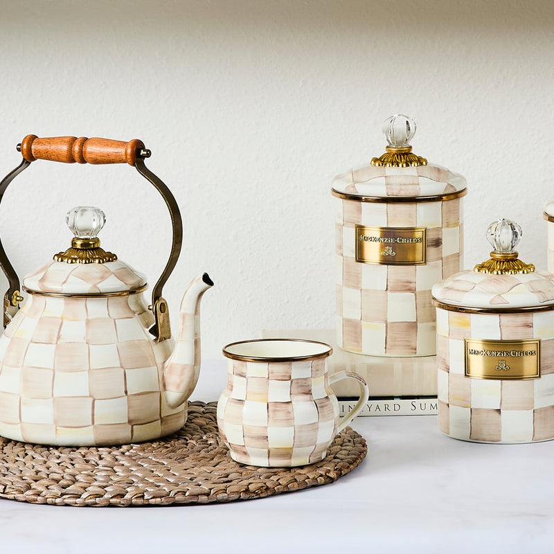 MacKenzie Childs | Mocha Check Mug showcased on a woven placemat, featuring hand-painted checks and bronzed stainless steel rim, with matching ceramic tea set and canisters.