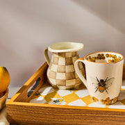 The MacKenzie Childs Mocha Check Creamer, featuring cream-and-tan checks, sits on a bee-patterned tray, ideal for versatile kitchen use.