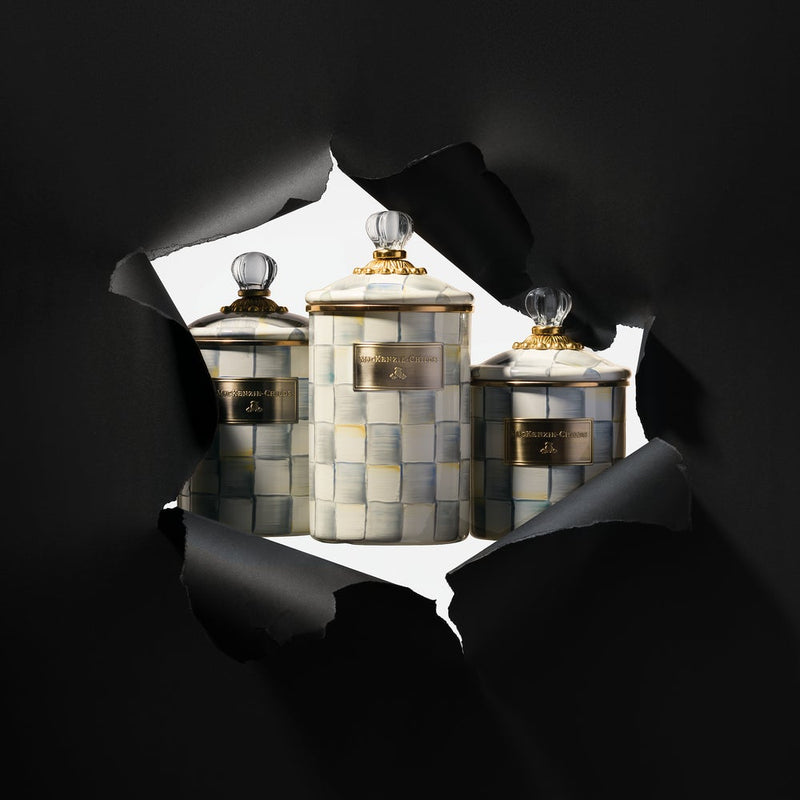 MacKenzie Childs | Sterling Check Large Canister trio with hand-painted checks, gold-tone bands, and clear glass knob lids, displayed through torn black paper.