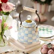 MacKenzie-Childs Sky Check Tea Kettle, 2 Qt, with a blue-and-white checkered pattern, sits on books amidst pink flowers and pastel tableware.