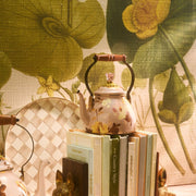 MacKenzie-Childs Wild Rose Mauve Tea Kettle, 2 Qt, sits elegantly on stacked books with a floral backdrop, featuring a bronzed rim and acacia handle.