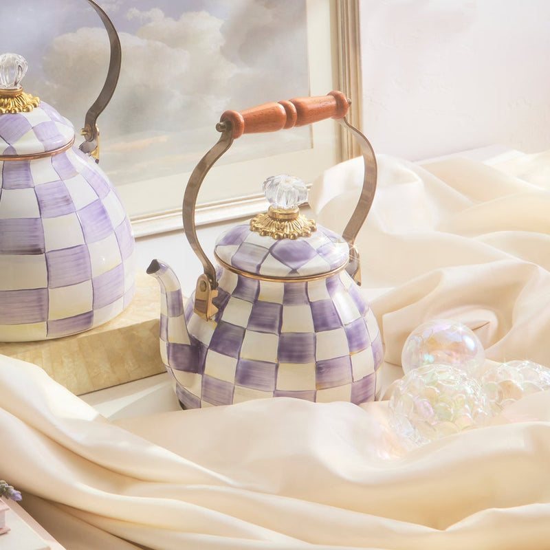 MacKenzie-Childs Violet Check Tea Kettle, 2 Qt, featuring a lavender checkerboard pattern, brass fittings, and a wood handle, elegantly displayed on ivory fabric.