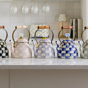 MacKenzie-Childs Violet Check Tea Kettle, 2 Qt, sits elegantly on a white countertop, surrounded by matching checkered kitchenware, showcasing its distinctive hand-painted design.