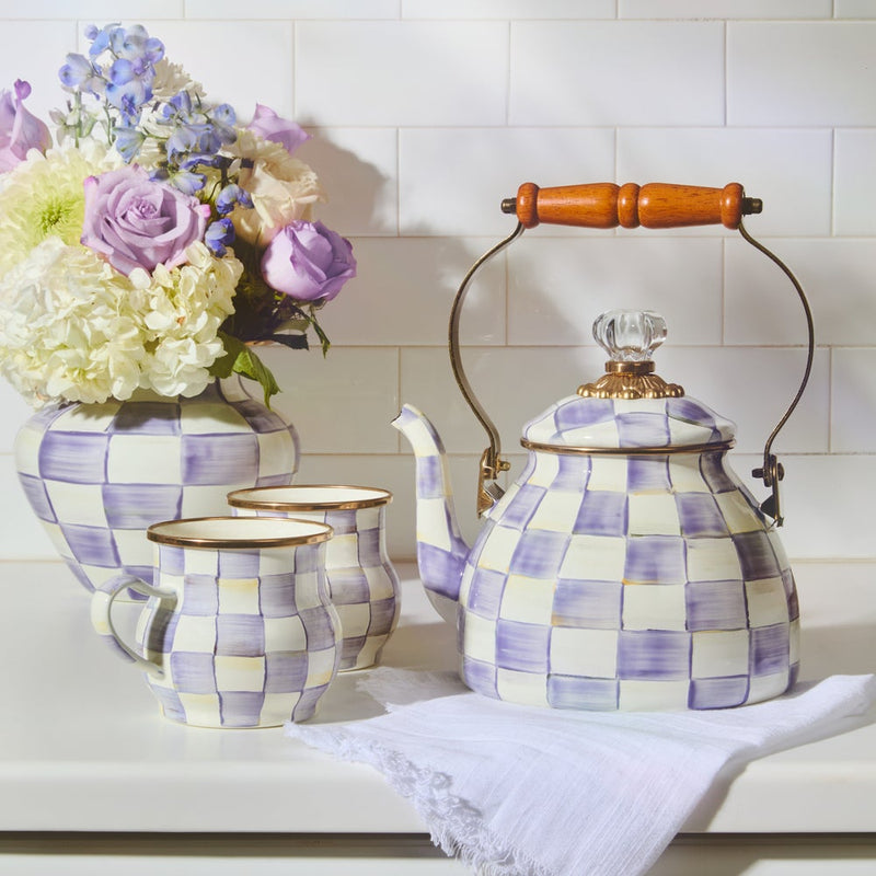 MacKenzie-Childs Violet Check Tea Kettle, 2 Qt, with wooden handle and crystal knob, sits elegantly on a countertop, flanked by matching mugs and a floral vase.