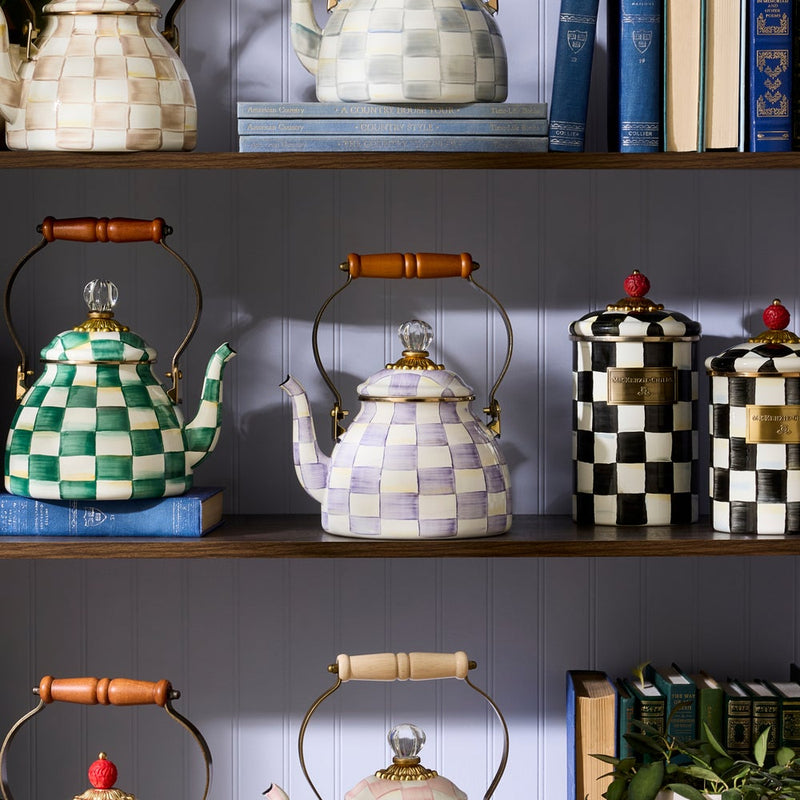 MacKenzie-Childs Violet Check Tea Kettle, 2 Qt: A hand-painted purple checkered kettle with brass accents and wooden handle, displayed on a shelf with matching canisters and books.