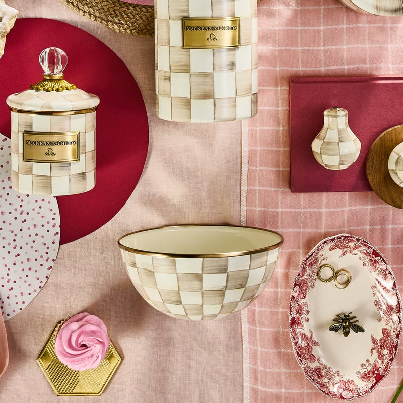 MacKenzie-Childs Mocha Check Small Everyday Bowl amid hand-painted ceramic canisters, a check-painted shaker, cupcake on gold plate, and decorative dish, all on pink fabric.