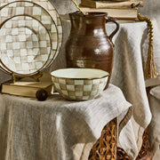 MacKenzie Childs | Mocha Check Small Everyday Bowl displayed in a rustic setting with matching ceramics, an earthenware jug, and vintage books on a wooden stool.