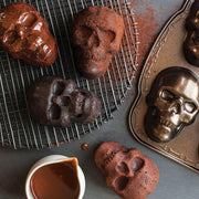 Nordic Ware | Skull Cakelet Pans - Skull Bites with skull-shaped cakes on a cooling rack, chocolate ganache, cocoa dust, and a pitcher of sauce for Halloween baking.