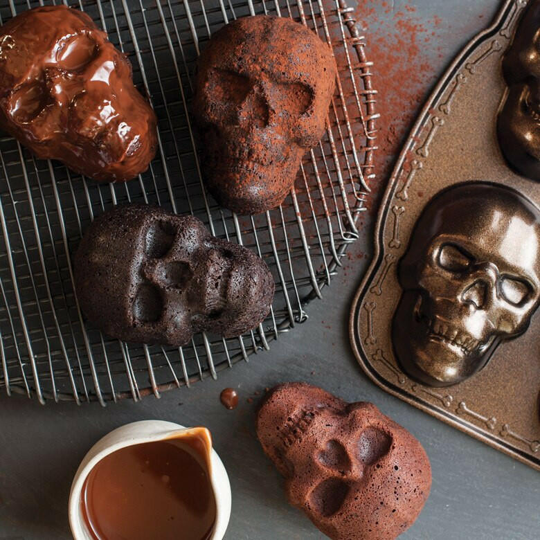 Nordic Ware | Skull Cakelet Pans - Skull Bites with skull-shaped cakes on a cooling rack, chocolate ganache, cocoa dust, and a pitcher of sauce for Halloween baking.