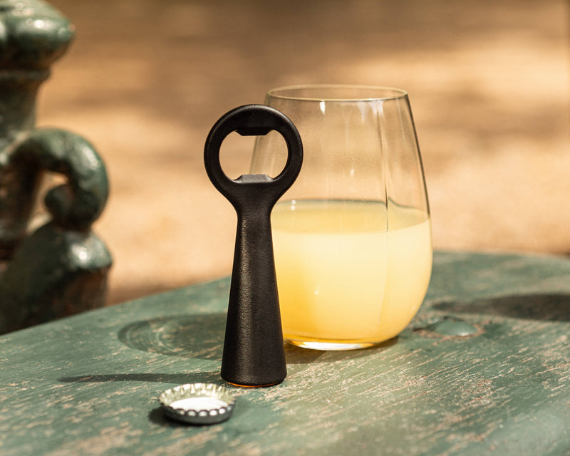 L'Atelier du Vin - USA - Bottle Opener - LE BRASSEUR – BLACK stands upright beside a tumbling glass, showcasing its sleek, stainless steel design on a weathered table.
