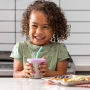 Young girl holding a PlanetBox 7oz Stainless Steel Cup with straw, smiling at a kitchen counter with a breakfast tray of waffles and fruit slices.