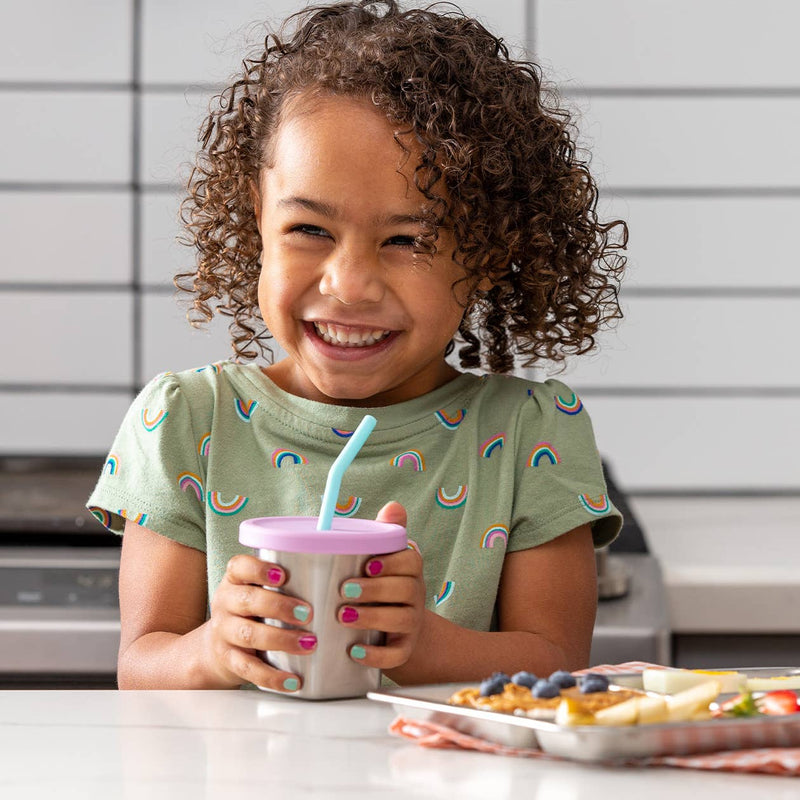 Young girl holding a PlanetBox 7oz Stainless Steel Cup with straw, smiling at a kitchen counter with a breakfast tray of waffles and fruit slices.