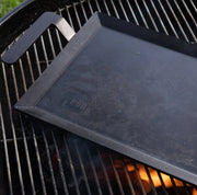 Made In | Carbon Steel Griddle on kettle-style charcoal grill, showing its round grate, glowing coals, and ergonomic handle, perfect for diverse cooktops.