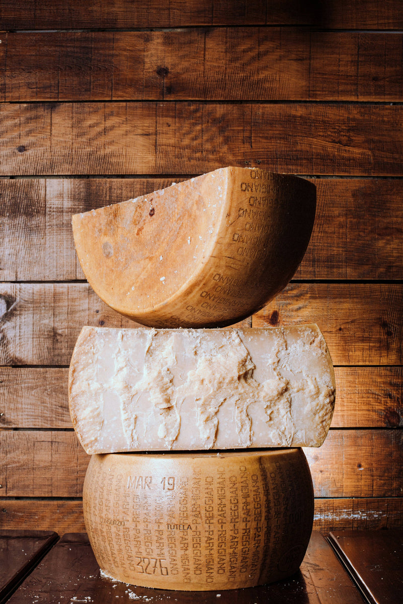 Nicola's Marketplace - Parmigiano Reggiano DOP Mitica® 36 months: 7 Ounces. Stacked aged cheese wheels, revealing crumbly interior and stamped rind, showcasing artisanal quality.