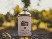 Pink House Alchemy - Vanilla Bean Simple Syrup bottle on a rock, featuring a label with a house illustration and a red circle with white numbers.