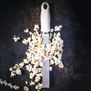 Microplane Premium Zester, Grater - Cashmere Beige, surrounded by popcorn, showcasing its fine rasp design on a dark surface, ideal for zesting and grating.