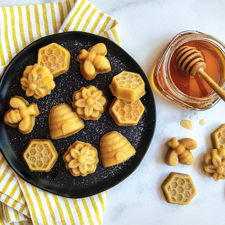 Nordic Ware Busy Bee Bitelet Pan featuring 16 cavities for creating bite-sized treats, shown with a plate of cookies and a honey dipper jar nearby.