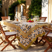 Couleur Nature - Forest Harvest Mustard & Blue tablecloth elegantly drapes a wooden table, set with wine, fruit, and cheese, in a sunlit garden dining scene.