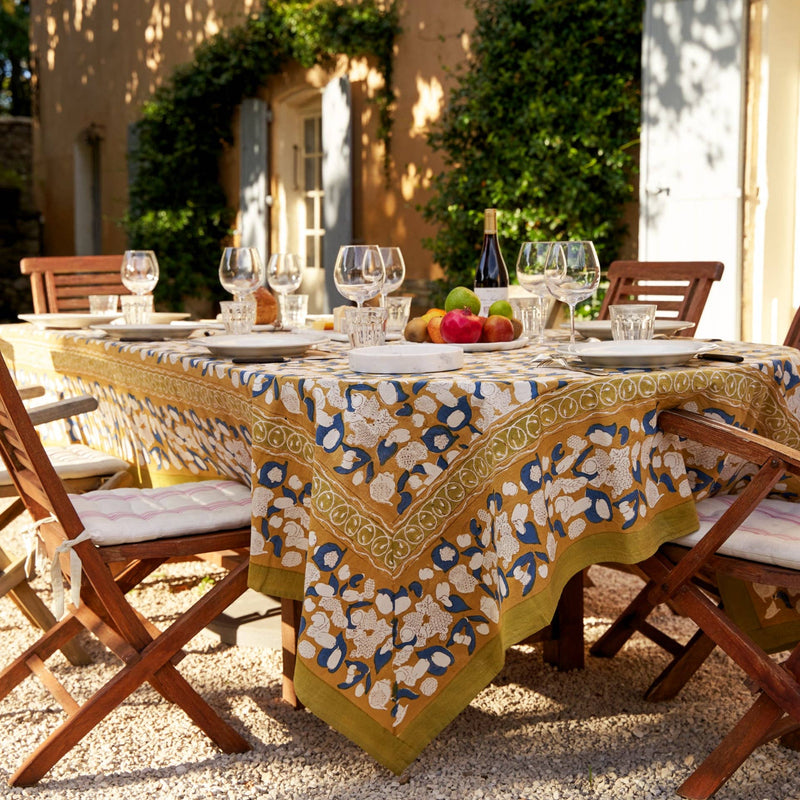 Couleur Nature - Forest Harvest Mustard & Blue tablecloth elegantly drapes a wooden table, set with wine, fruit, and cheese, in a sunlit garden dining scene.