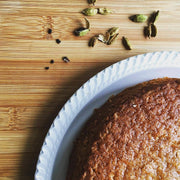 Golden-brown cardamom cake slice on white plate with scattered green cardamom pods, showcasing Burlap & Barrel - Cloud Forest Cardamom - Single Origin Spice & Seasoning: 1.7 oz glass jar.