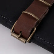 Close-up of Messermeister Vintage Leather 7 Pocket Knife Roll - Black, featuring a dark brown leather strap with antiqued brass buckle, stitched on a black background.