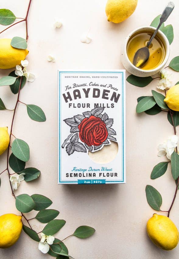 Hayden Flour Mills Semolina Flour surrounded by lemons, green foliage, white blossoms, and a cup of oil, styled on a light surface. Perfect for pasta and baking.