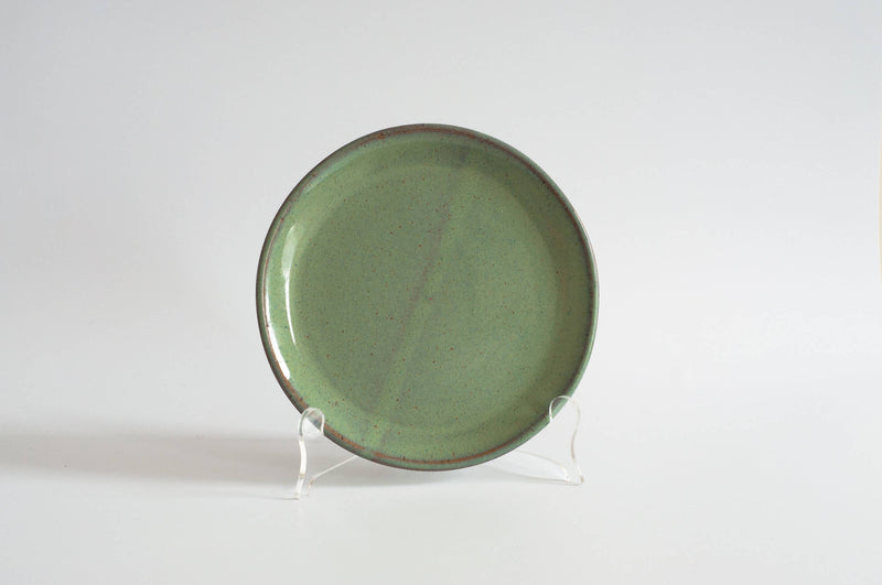RachaelPots - Salad Plate: Coral, a handcrafted round green-glazed ceramic plate with brown speckles, displayed on a clear stand, highlighting its slightly upturned lip and flat center.