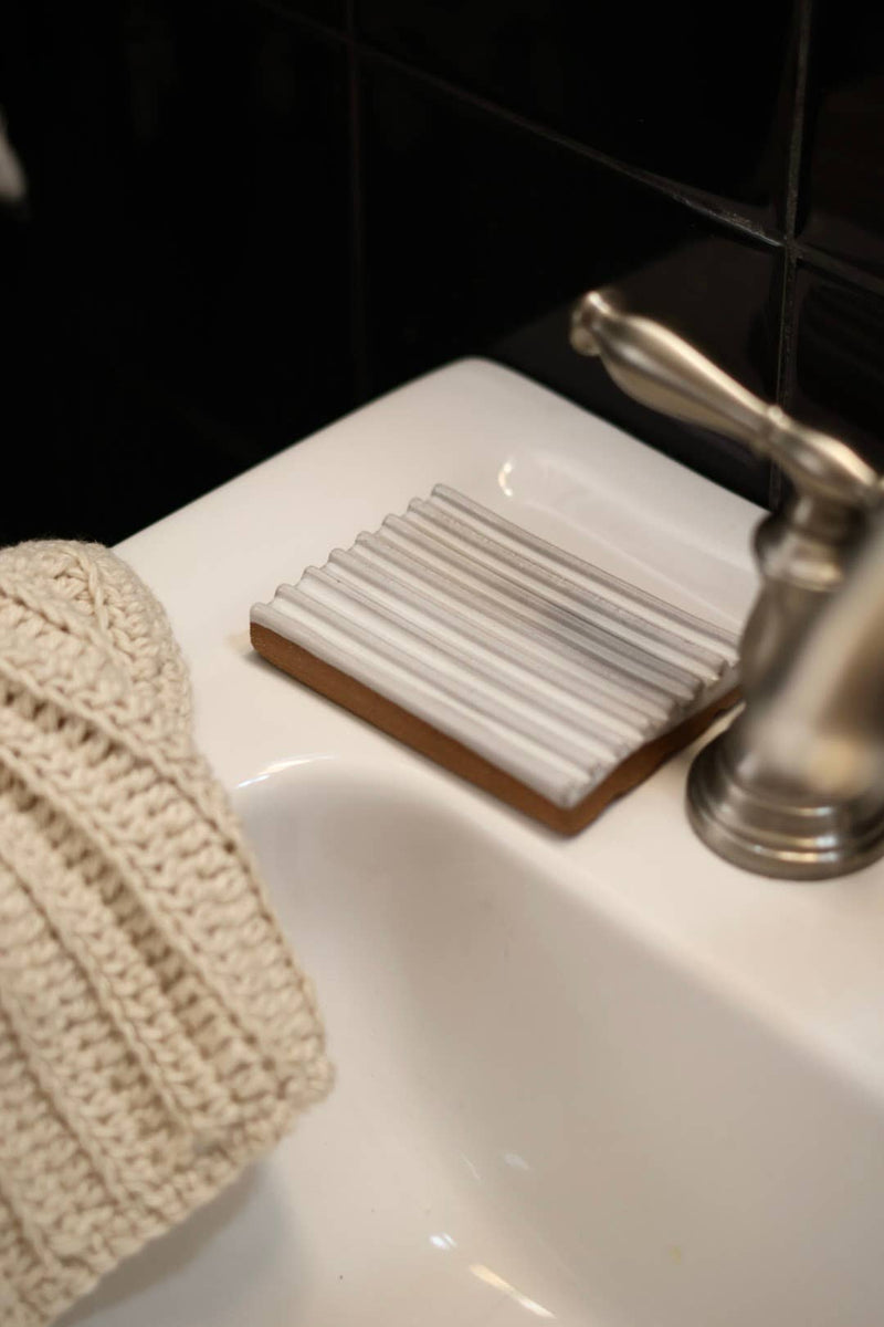 Gravesco Pottery - Soap Dish | White: Close-up of a handmade ceramic soap dish on a wooden base, showcasing its unique ridged design beside a satin-nickel faucet in a bathroom setting.