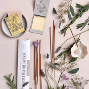 The Floral Society Essential Oil Incense set with lavender, lemon eucalyptus, and rosemary, featuring incense sticks, ceramic holder, matches, and botanical accents on a pink surface.