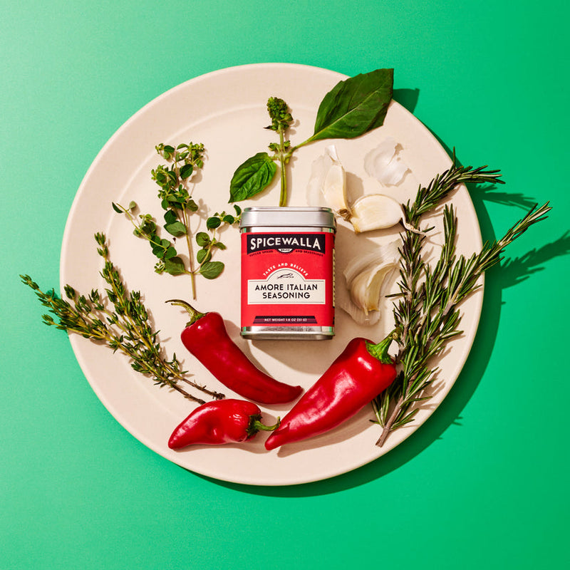 Spicewalla Amore Italian Seasoning tin surrounded by chili peppers, garlic, rosemary, thyme, and basil on a plate, highlighting its use in Italian cooking.