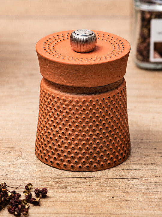 Peugeot | Bali Cast Iron Pepper Mill, Orange, on a wooden surface with peppercorns and a spice jar, highlighting its unique dimpled design inspired by traditional tea ceremonies.