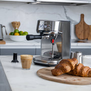 Breville | Bambino Plus espresso machine on marble countertop with latte, tamper, milk pitcher, and croissants, showcasing professional cafe quality in a kitchen setting.