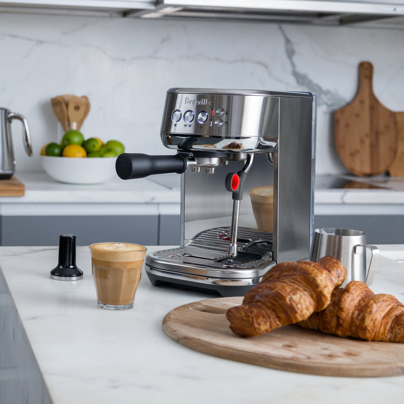 Breville | Bambino Plus espresso machine on marble countertop with latte, tamper, milk pitcher, and croissants, showcasing professional cafe quality in a kitchen setting.