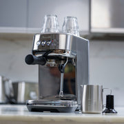 Breville | Bambino Plus espresso machine on kitchen counter, with shot glasses, portafilter, steam wand, milk frothing pitcher, and tamper; ideal for authentic coffee creation.