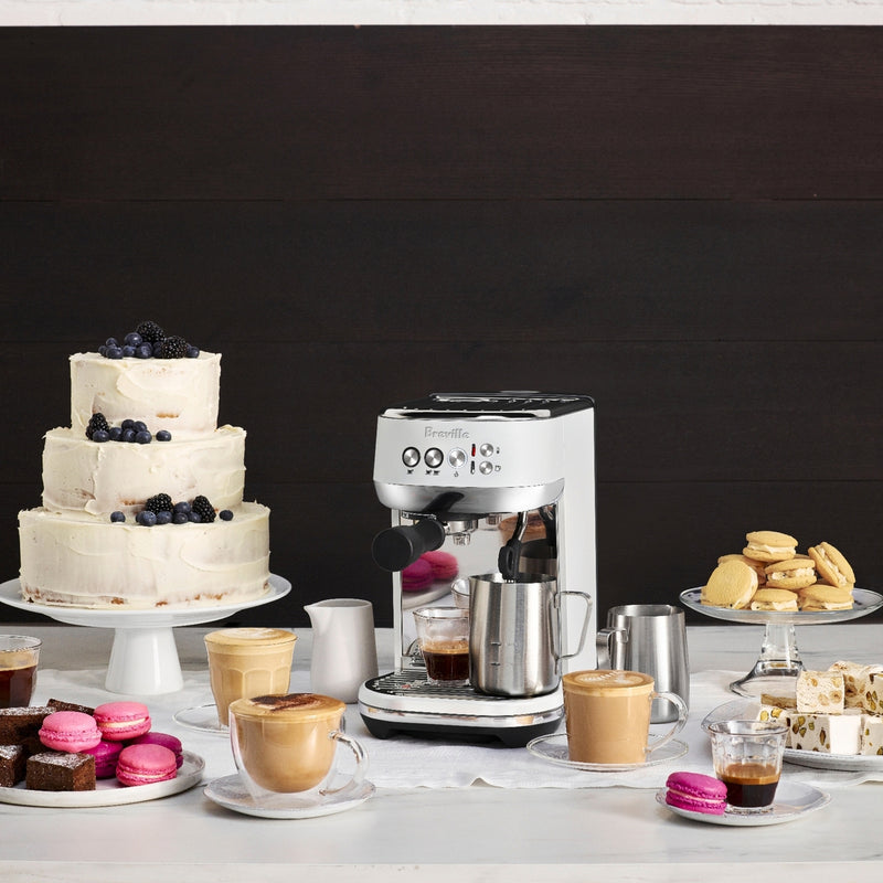 Breville | Bambino Plus espresso machine with lattes, espresso, and assorted desserts, including a tiered cake and macarons, showcasing Athens Cooks' curated kitchen selection.