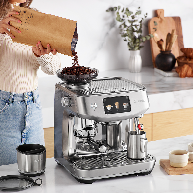 Breville | The Oracle Jet, Black Truffle espresso machine with beans being poured into the hopper, surrounded by a barista setup including a knock-box and milk pitcher.
