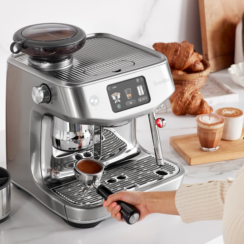 Breville | The Oracle Jet, Black Truffle espresso machine with touchscreen and grinder, on a marble countertop, portafilter held, croissants and latte art drinks in background.