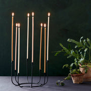 The Floral Society | Long Stem Beeswax Candles, Ochre, elegantly flicker in a black-metal candelabra, creating a warm ambiance alongside leafy houseplants on a simple tabletop.