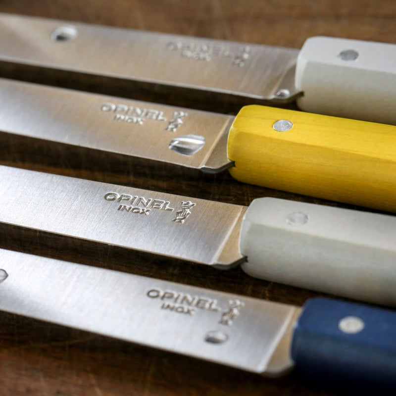 Opinel | Bon Appétit Steak Knives, Navy Blue, featuring sharp stainless steel blades and colorful wood handles, laid on a wooden surface.