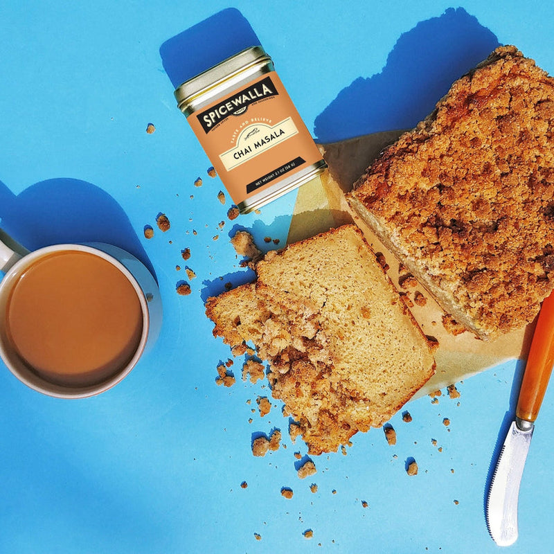 Spicewalla Chai Masala tin with a mug of chai, crumb-topped loaf slices, and an orange-handled knife; perfect for enhancing your chai experience.