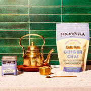 Spicewalla | Chai Pani Ginger Chai in tin and resealable bag, with a brass teapot, cup of cardamom pods, and teaspoon, on a speckled countertop.