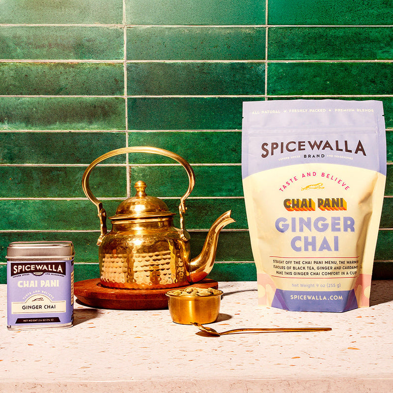 Spicewalla | Chai Pani Ginger Chai setup featuring a tin, brass kettle, cardamom bowl, spoon, and a large pouch on a countertop against green tiles.