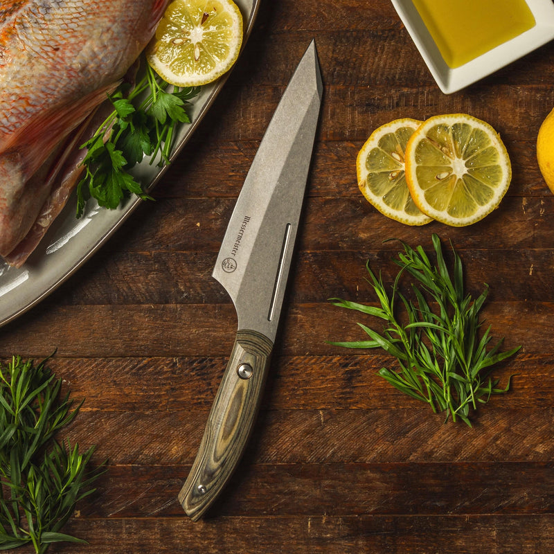 Messermeister | Chef April Bloomfield Chef's Knife, 6.5 inches on a wooden surface with herbs, lemon, and olive oil, ideal for precise kitchen prep.