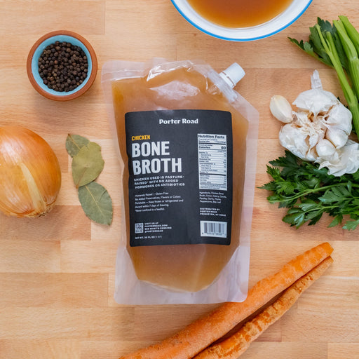 Porter Road Butcher | Chicken Bone Broth with fresh vegetables, garlic, and herbs on a table, ideal for enhancing soups and stews.
